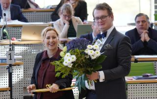 Franziska Schubert und Valentin Lippmann stehen im Plenarsaal. Franziska hält ein Holzbrett und Valentin einen Blumenstrauß.